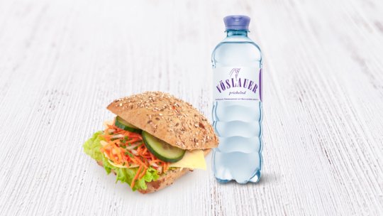 Ein Mehrkornsandwich mit Salat, Käse, geraspelten Karotten und Gurkenscheiben neben einer blauen Flasche Vöslauer Mineralwasser auf einer weißen Holzfläche.