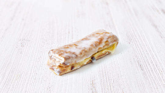 Ein rechteckiges Gebäck mit glasiertem Zuckerguss, gefüllt mit einer gelben Creme und einer einzelnen Heidelbeere, auf einer weißen Holzunterlage.