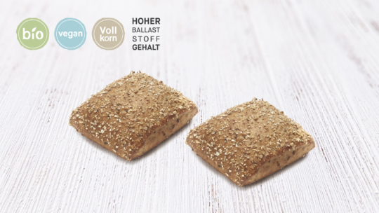 Zwei quadratische Mehrkornbrötchen auf einer weißen Holzunterlage. Auf dem Etikett steht: bio, vegan, Vollkorn und HOHER BALLASTSTOFFGEHALT auf Deutsch, was auf Bio, vegan, Vollkorn und hohen Ballaststoffgehalt hinweist.