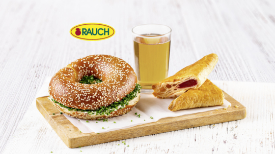 Ein Sesambrötchen mit grünen Kräutern, ein Glas Apfelsaft und ein mit Früchten gefülltes Gebäck sind auf einem Holzbrett mit dem Rauch-Logo im Hintergrund angeordnet.