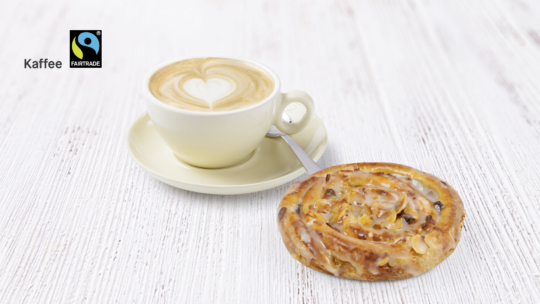 Eine Tasse Cappuccino mit herzförmigem Schaum und einem Gebäckstück auf einer weißen Holzfläche. Das Fairtrade-Logo und das Wort Kaffee erscheinen in der oberen linken Ecke.