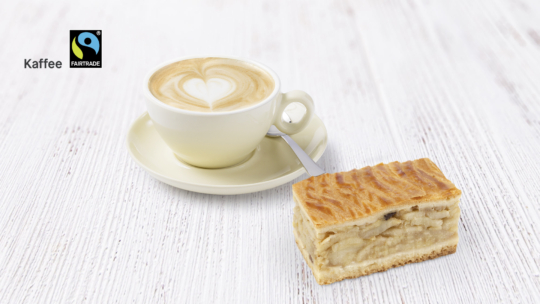 Eine Tasse Cappuccino mit Latte Art steht auf einer Untertasse neben einem Stück Apfelkuchen auf einer hellen Holzfläche. Das Fairtrade-Logo und das Wort Kaffee erscheinen in der oberen linken Ecke.