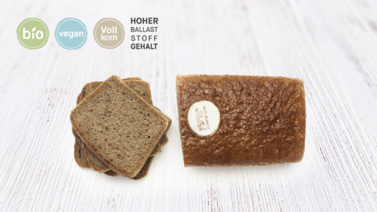 Ein Laib dunkles Vollkornbrot, teilweise aufgeschnitten, auf einer weißen Holzfläche. Die vier Symbole darüber lauten: bio, vegan, Vollkorn und hoher Ballaststoffgehalt.