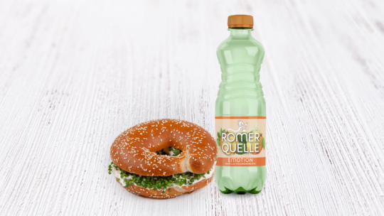Ein Sesambrötchen mit Frischkäse und Kräutern liegt neben einer grünen Plastikflasche Römerquelle Emotion auf einer hellen Holzfläche.