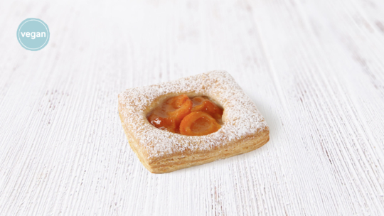 Ein quadratisches Fruchtgebäck mit Puderzucker und Orangenscheiben liegt auf einer weißen Holzfläche. In der oberen linken Ecke befindet sich ein türkisfarbenes veganes Etikett.