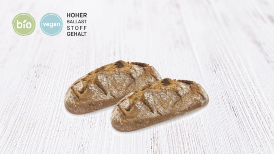 Zwei rustikale Brotlaibe auf einer weißen Holzfläche. In der oberen linken Ecke stehen runde Etiketten mit der Aufschrift "Bio" und "Vegan", daneben die Aufschrift "HOHER BALLASTSTOFFGEHALT".