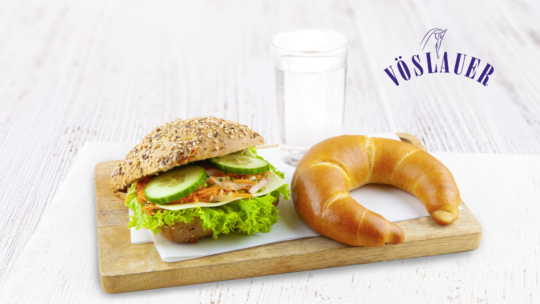 Ein belegtes Sandwich mit Salat, Gurke und Gemüse, ein Croissant und ein Glas Wasser stehen auf einem Holzbrett. Im Hintergrund ist das Vöslauer-Logo zu sehen.