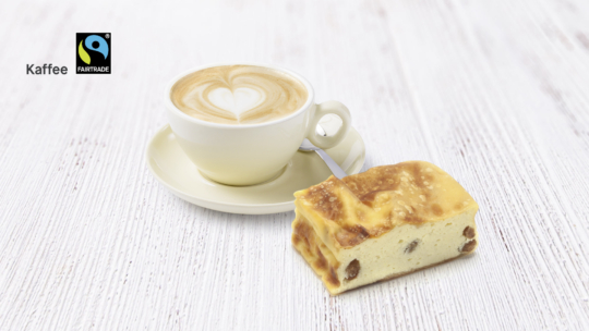 Eine Tasse Cappuccino mit herzförmigem Schaum sitzt auf einer Untertasse mit einem Löffel, daneben ein Stück Käsekuchen mit Rosinen. Der Hintergrund ist eine helle Holzoberfläche. In der Ecke erscheinen das Fairtrade-Logo und das Wort Kaffee.