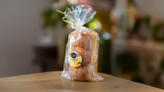 Ein Laib Brot ist in einer durchsichtigen, oben verschlossenen Plastiktüte verpackt, die auf einer Holzunterlage steht. Auf der Verpackung befindet sich ein gelbes Etikett mit Text. Der Hintergrund ist sanft verschwommen.