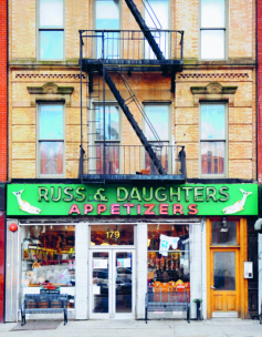 Ein Schaufenster mit einem grünen Schild mit der Aufschrift Russ & Daughters Appetizers in weißen Buchstaben und Abbildungen von Fischen. Im Schaufenster sind verschiedene Waren ausgestellt, und über dem Eingang des Geschäfts befindet sich eine Feuerleiter.