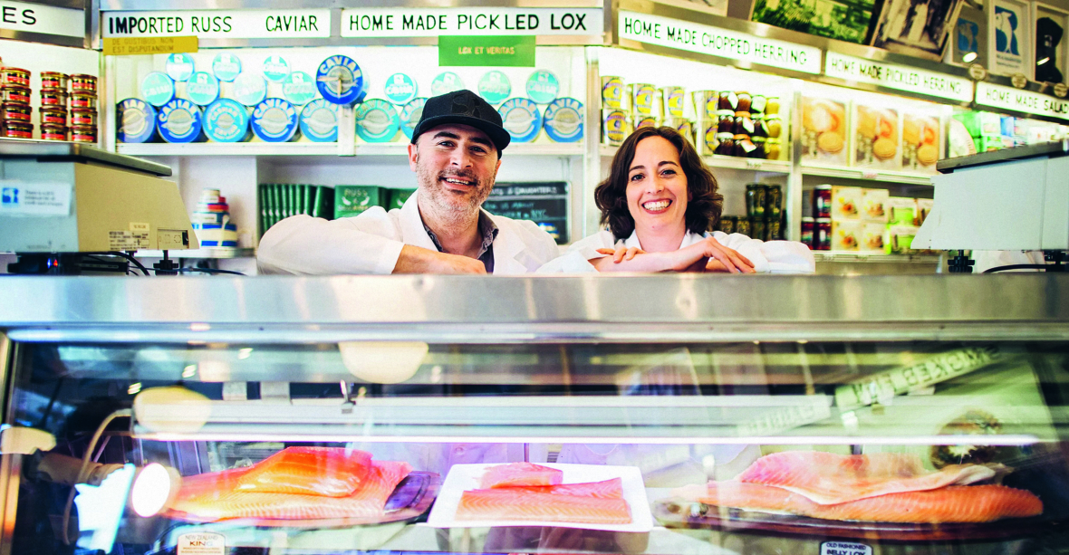 Zwei Personen in weißen Kitteln stehen lächelnd hinter einer Fischtheke, die Fischfilets ausstellt. Hinter ihnen stehen Regale mit Konserven und Schildern, die für Kaviar, eingelegten Lachs und andere Delikatessen werben.