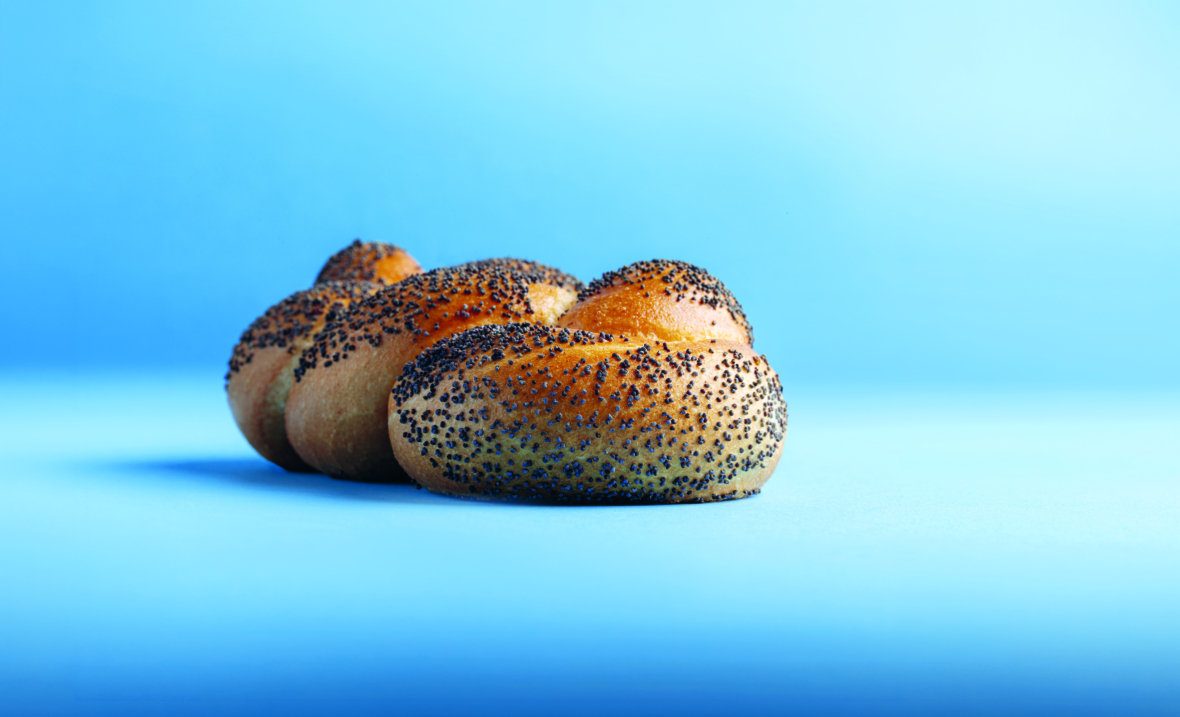 Ein goldbraunes, geflochtenes Brötchen mit schwarzem Mohn, fotografiert vor einem hellblauen Hintergrund mit Farbverlauf.