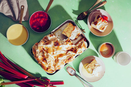 Auf einem grünen Tisch steht ein Tablett mit gebackener Alaska mit goldenem Baiser, die Scheiben werden auf Tellern serviert. Daneben stehen Tassen, Gabeln, ein Topf mit Pudding, eine Schüssel mit Rhabarbersauce und frische Rhabarberstangen.