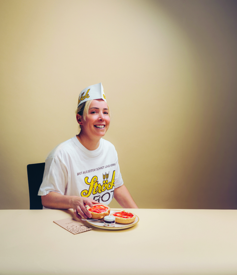 Eine lächelnde Person mit einem weißen Papierhut und einem Ströck-T-Shirt sitzt an einem Tisch mit zwei belegten Brötchen mit Tomatenscheiben und einem kleinen Behälter mit Aufstrich.