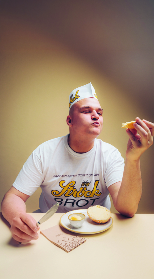 Eine Person mit einer weißen Bäckermütze und einem T-Shirt sitzt an einem Tisch, lächelt und hält ein Stück Brot in der Hand. Auf dem Tisch liegen ein Messer, Butter und noch mehr Brot. Der Hintergrund ist schlicht und sanft beleuchtet.