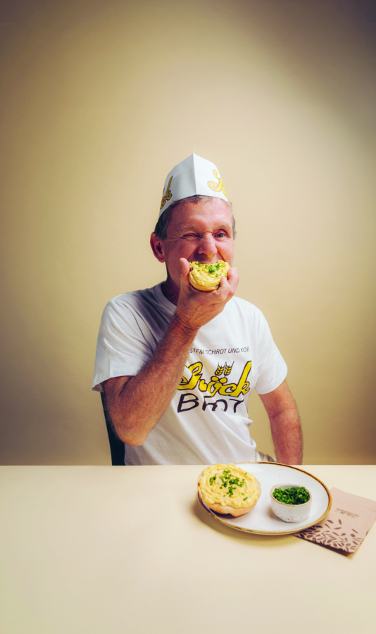 Ein älterer Mann mit weißem Hut und T-Shirt sitzt an einem Tisch und isst lächelnd ein Gebäckstück. Vor ihm stehen ein Teller mit einem weiteren Gebäckstück, eine kleine Schale mit grünem Dip und eine braune Serviette.