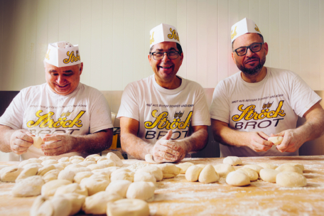 Drei lächelnde Bäcker in weißen T-Shirts und Papierhüten der Marke Ströck Brot formen auf einem mit Mehl bestäubten Holztisch in einer Bäckerei Brötchen aus Brotteig.