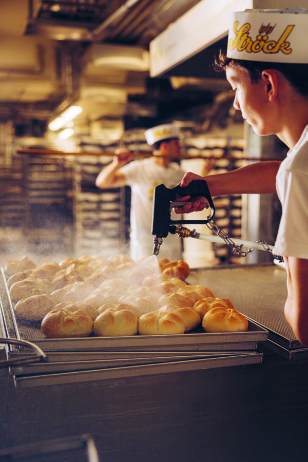 Ein Bäcker in weißer Uniform und mit Mütze sprüht in einer Bäckerei Wasser auf die Bleche mit frisch gebackenen Brötchen, während ein anderer Bäcker im Hintergrund zwischen den Brotständern arbeitet.
