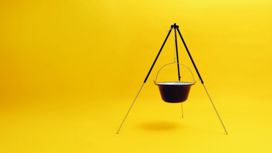 Ein schwarzer Kochtopf hängt an einem Stativständer vor einem leuchtend gelben Hintergrund. Der Aufbau ist minimalistisch, mit Stativ und Topf in der Mitte und viel freiem Raum um sie herum.