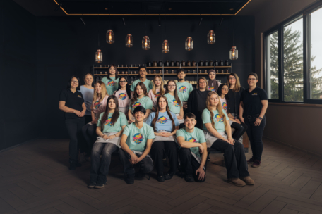 Eine Gruppe von zwanzig Personen, zumeist junge Erwachsene in passenden hellblauen T-Shirts mit einem bunten Logo, posiert in einem modernen Raum mit Holzfußboden, Regalen und Hängelampen. Zwei Erwachsene in schwarzen Hemden stehen an den Seiten.