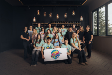 Eine Gruppe Jugendlicher und Erwachsener posiert in einem Innenraum und trägt passende bunte T-Shirts. Zwei halten ein großes Schild mit der Aufschrift "Laboratório". Hinter ihnen stehen Flaschen in Regalen, und von der Decke hängen Pendelleuchten.