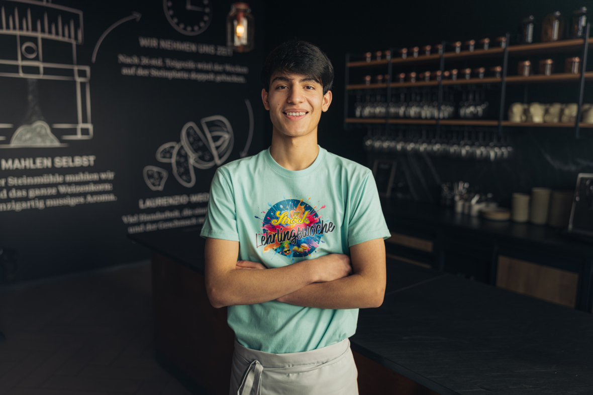 Ein junger Mann mit dunklem Haar, der ein buntes T-Shirt und eine helle Schürze trägt, steht lächelnd mit verschränkten Armen in einem modernen, schwach beleuchteten Café mit Regalen und Kreidekunst an den Wänden im Hintergrund.