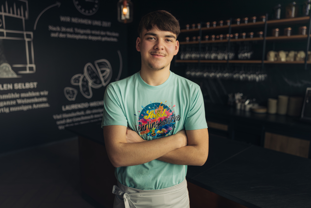 Ein junger Mann mit kurzen braunen Haaren steht selbstbewusst mit verschränkten Armen, einem bunten T-Shirt und einer weißen Schürze in einer modernen, schwach beleuchteten Küche mit Regalen und einer Kreidetafel im Hintergrund.