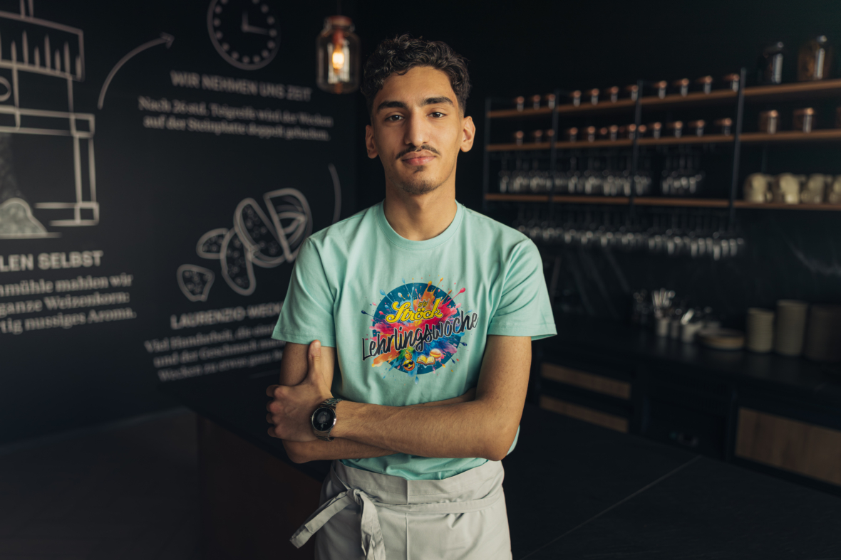 Ein junger Mann mit kurzen lockigen Haaren und einem Schnurrbart steht selbstbewusst in einer modernen Küche, die Arme verschränkt. Er trägt ein hellblaues Hemd mit bunten Grafiken und eine weiße Schürze, im Hintergrund sind Regale und Küchenutensilien zu sehen.
