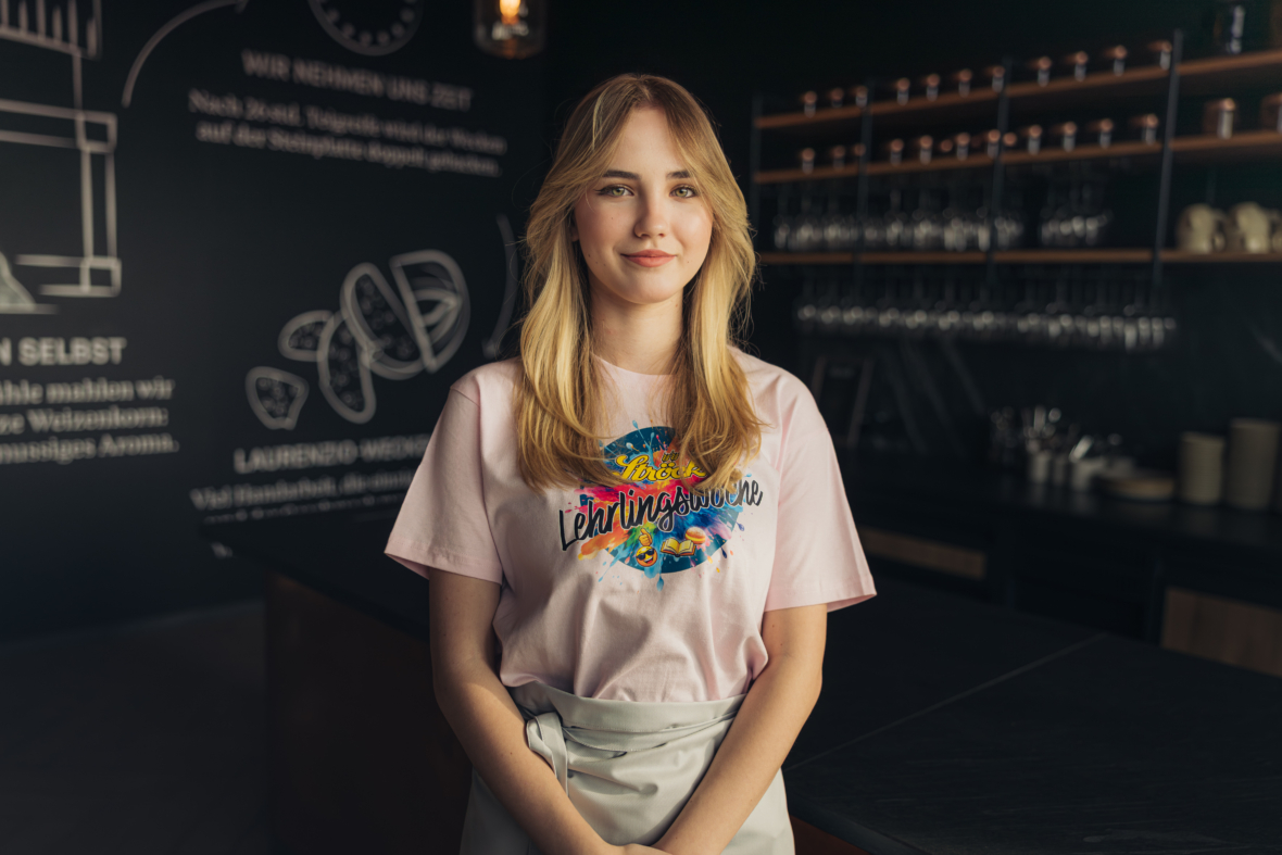 Eine junge Frau mit langen blonden Haaren steht in einer modernen Küche und trägt ein blassrosa T-Shirt mit einer bunten Grafik und eine helle Schürze. Im Hintergrund sind Regale mit Glaswaren und eine Kreidetafel zu sehen.