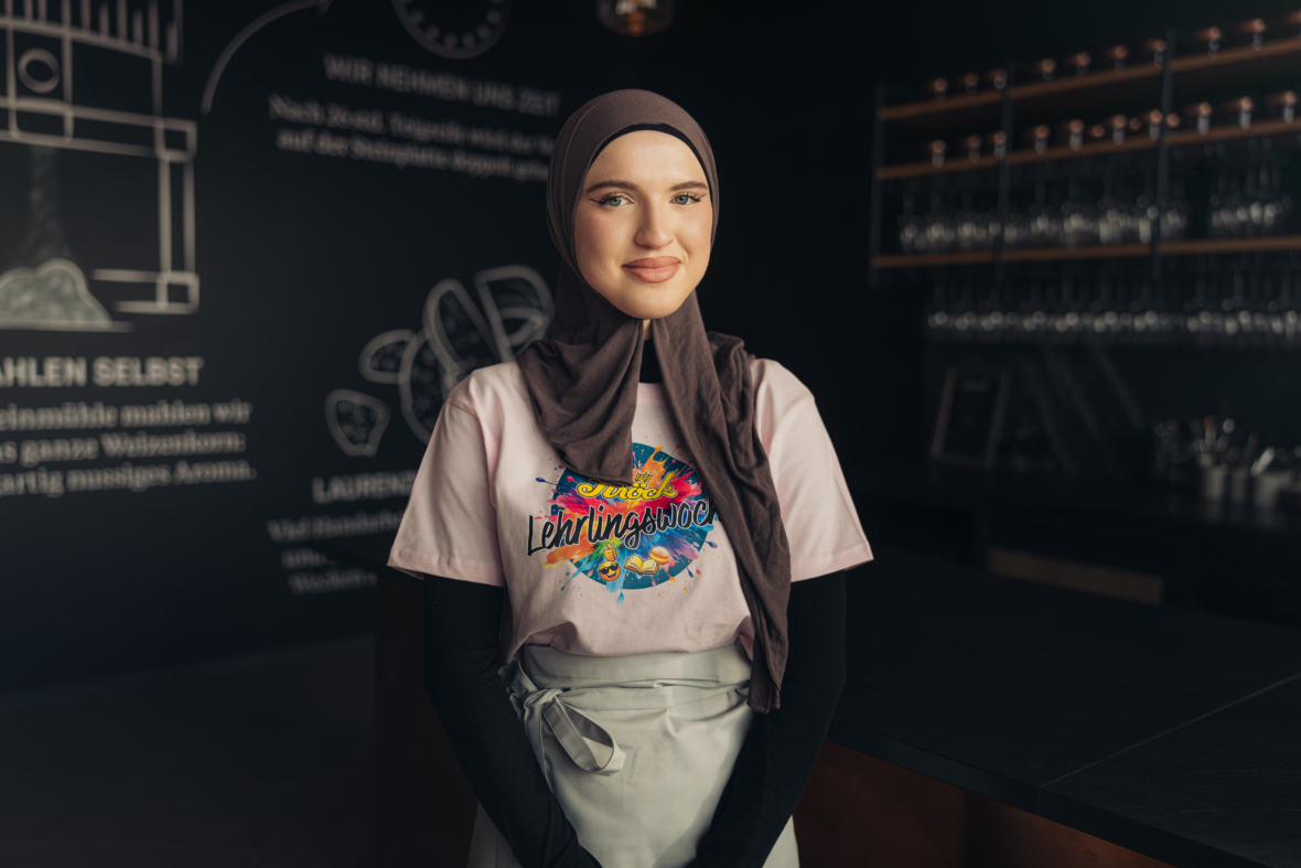Eine junge Frau, die einen Hidschab, ein buntes Lehrlingshelden-T-Shirt und eine Schürze trägt, steht lächelnd in einem modernen, schwach beleuchteten Restaurant oder Café mit Regalen und Kreidezeichnungen im Hintergrund.