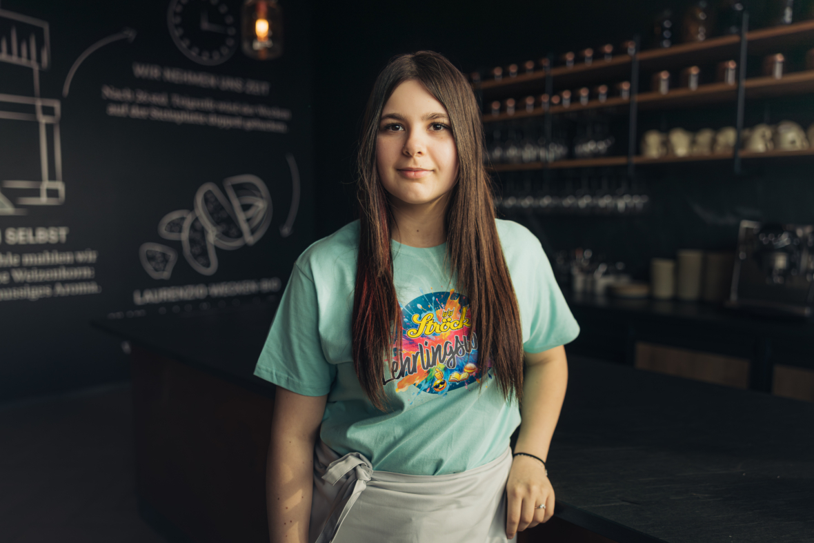 Eine junge Frau mit langen braunen Haaren steht in einem modernen Café, trägt ein buntes T-Shirt und eine weiße Schürze, im Hintergrund sind Regale mit Tassen und eine Speisekarte auf einer Kreidetafel zu sehen.