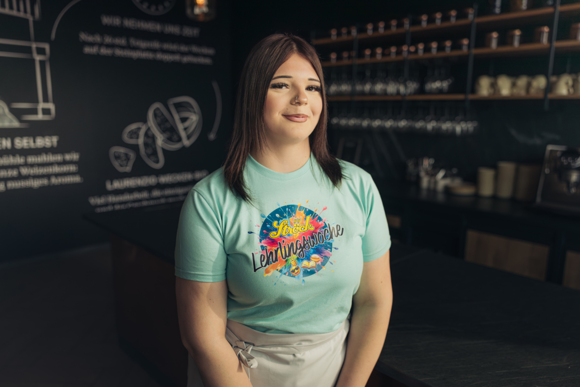 Eine junge Frau mit glattem braunem Haar, die ein buntes Lerningshelden-T-Shirt und eine weiße Schürze trägt, steht lächelnd in einem modernen, schwach beleuchteten Café mit Regalen voller Tassen und einer Kreidetafel hinter ihr.