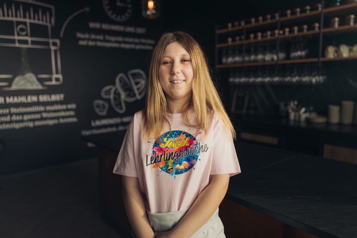 Eine junge Frau mit langen, hellbraunen Haaren steht lächelnd in einer modernen Küche. Sie trägt ein hellrosa T-Shirt mit einer bunten Grafik, auf der "Lieblingsbäcker" steht. Im Hintergrund sind Regale mit Gläsern und Küchenutensilien zu sehen.
