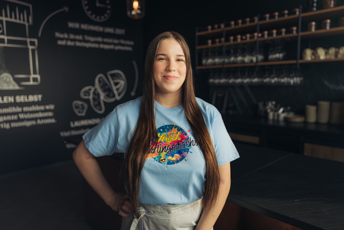Eine junge Frau mit langen braunen Haaren steht lächelnd in einem modernen Café, trägt ein hellblaues T-Shirt mit einer bunten Grafik und eine Schürze. Hinter ihr stehen Regale mit Gläsern und Tellern und eine dunkle Wand mit Kreidezeichnungen.