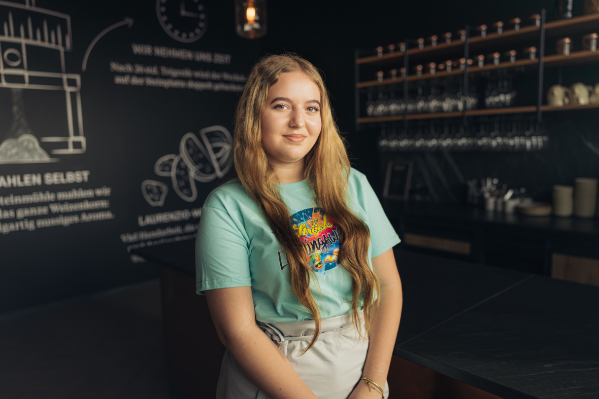 Eine junge Frau mit langem, gewelltem Haar steht lächelnd in einer modernen Küche mit dunklen Wänden und Regalen. Sie trägt ein hellgrünes grafisches T-Shirt und eine cremefarbene Hose. Die Wand hinter ihr ist mit Bildern und Texten im Stil einer Kreidetafel verziert.