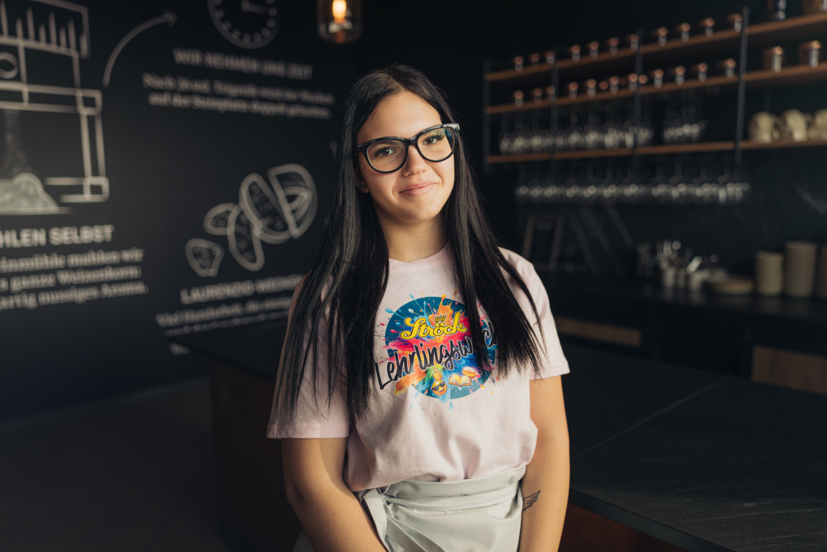 Eine junge Frau mit langen dunklen Haaren und Brille steht lächelnd in einer modernen Küche. Sie trägt ein hellrosa T-Shirt mit einer farbenfrohen Grafik und Text, mit Regalen und Küchengeräten im Hintergrund.