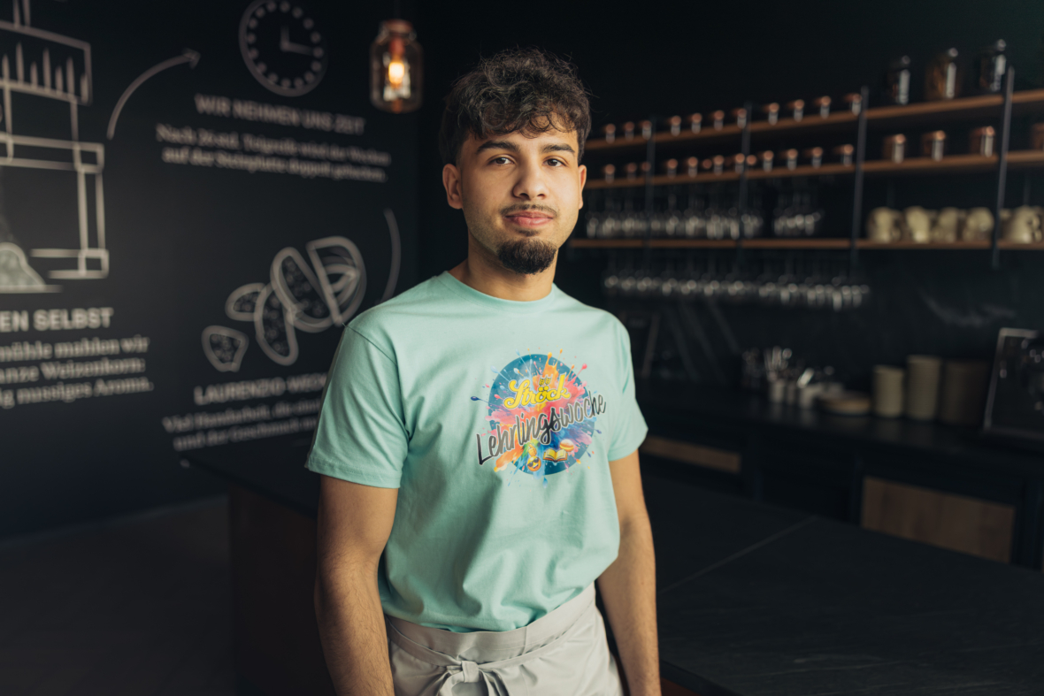 Ein junger Mann mit kurzen dunklen Haaren und Bart steht in einer modernen Caféküche, trägt ein buntes T-Shirt und eine helle Schürze. Im Hintergrund sind Regale mit Tassen und Gläsern sowie Wandmalereien im Stil einer Kreidetafel zu sehen.