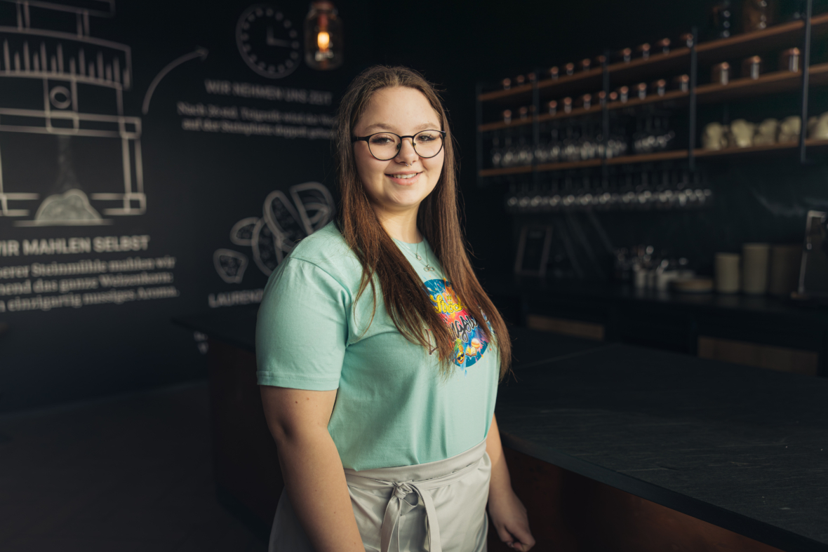 Eine junge Frau mit langen braunen Haaren, die eine Brille, ein hellblaues T-Shirt mit buntem Muster und eine Schürze trägt, steht lächelnd in einem modernen Café mit dunklen Wänden, Regalen und Glaswaren.