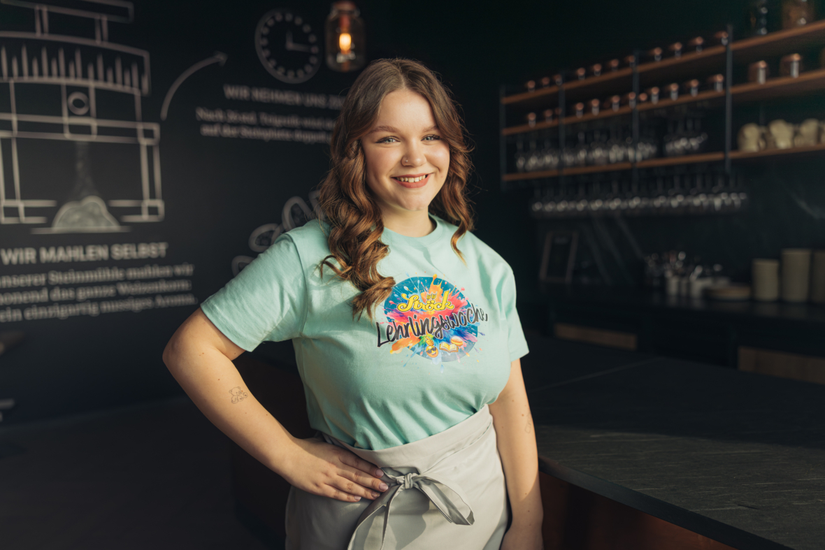 Eine junge Frau mit gewelltem braunem Haar steht lächelnd in einem modernen Café. Sie trägt ein hellblaues T-Shirt mit buntem Muster und eine graue Schürze und hat eine Hand in die Hüfte gestützt. Im Hintergrund befinden sich Regale mit Gläsern und eine Tafel.