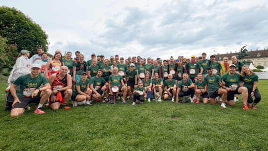 Eine große Gruppe von Läufern in zusammenpassenden grünen Hemden posiert vor einem Rennen gemeinsam auf einer Wiese und lächelt in die Kamera, während im Hintergrund Bäume und Gebäude unter einem bewölkten Himmel zu sehen sind.