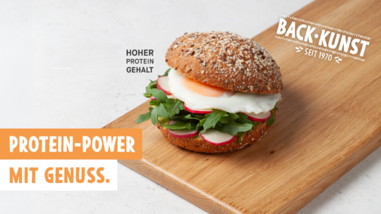 Ein Vollkornsandwichbrötchen mit Salat, Radieschenscheiben und einem pochierten Ei liegt auf einem Holzbrett. Der deutsche Text wirbt für den hohen Proteingehalt und den angenehmen Geschmack. BACK:KUNST SEIT 1970 erscheint oben rechts.