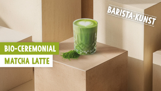 Ein Glas mit grünem Matcha-Latte und herzförmigem Schaum steht auf einem beigen Podest neben einem kleinen Haufen Matcha-Pulver. Der Text lautet BIO-CEREMONIAL MATCHA LATTE und BARISTA-KUNST.