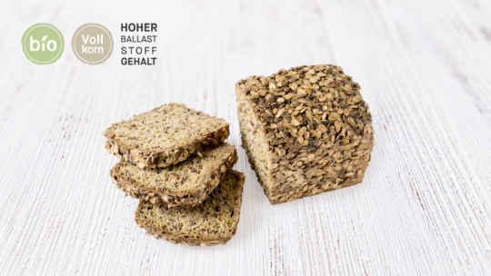 Ein Laib Vollkornbrot mit Haferflocken, teilweise aufgeschnitten, liegt auf einer weißen Holzfläche. Deutsche Etiketten weisen darauf hin, dass es biologisch, aus Vollkorn und reich an Ballaststoffen ist.