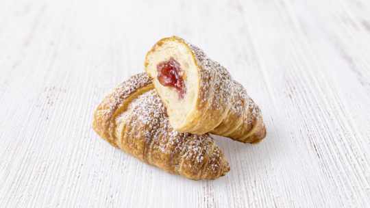 Zwei goldbraune, mit Puderzucker bestäubte Croissants, von denen eines aufgeschnitten ist und eine Füllung aus roter Fruchtmarmelade enthält, auf eine helle Holzunterlage gelegt.