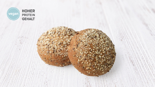 Zwei runde Vollkornbrötchen mit gemischten Körnern obenauf, auf einer weißen Holzunterlage. Auf dem Etikett steht auf Deutsch "vegan, hoher Proteingehalt".