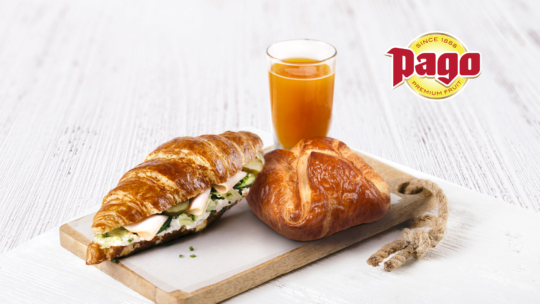 Ein Croissant-Sandwich mit Käse und Grünzeug, ein einfaches Croissant und ein Glas Orangensaft werden auf einem Holzbrett serviert. In der rechten oberen Ecke ist das Pago-Logo zu sehen.