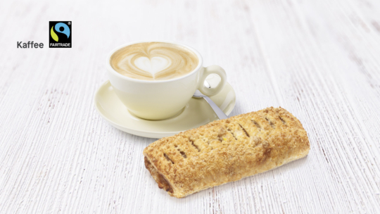 Eine Tasse Cappuccino mit herzförmiger Latte Art in einer Untertasse, ein Löffel und ein Blätterteiggebäck auf einer weißen Holzfläche. Das Fairtrade-Logo und das Wort Kaffee sind in der oberen linken Ecke zu sehen.