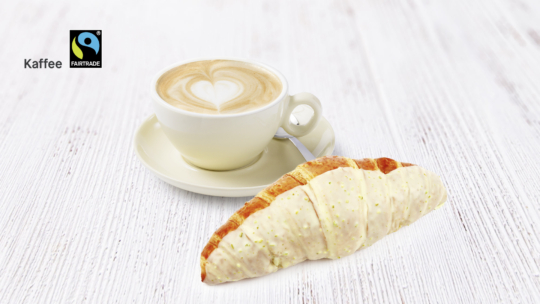 Eine Tasse Cappuccino mit herzförmigem Milchkaffee sitzt auf einer Untertasse neben einem leicht glasierten Croissant auf einer weißen Holzoberfläche. Das Fairtrade-Logo und das Wort Kaffee befinden sich in der oberen linken Ecke.