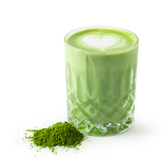Ein Glas mit grünem Matcha Latte mit herzförmiger Latte Art auf der Oberseite steht neben einem kleinen Haufen Matcha-Pulver auf weißem Hintergrund.