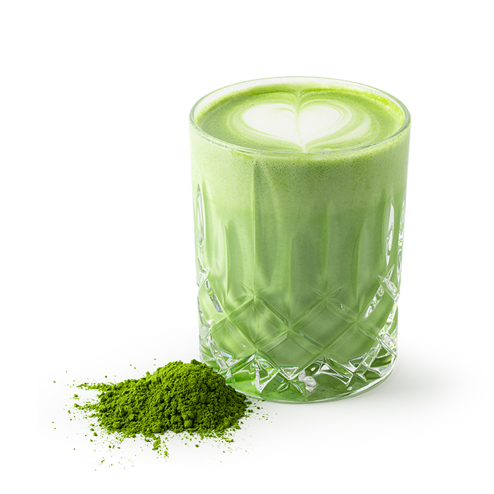 Ein Glas mit grünem Matcha Latte mit herzförmiger Latte Art auf der Oberseite steht neben einem kleinen Haufen Matcha-Pulver auf weißem Hintergrund.